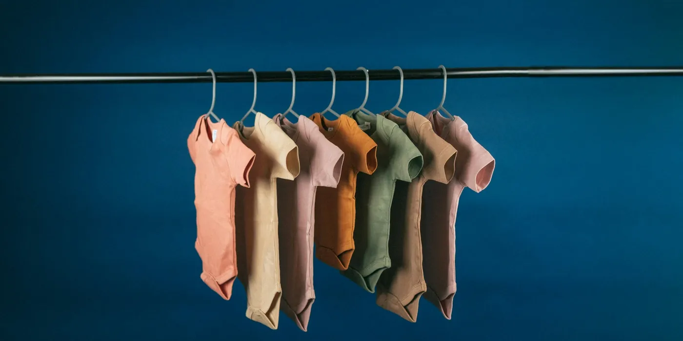 Row of tiny short sleeve baby bodysuits in soft neutrals hanging on a rail against a deep blue wall