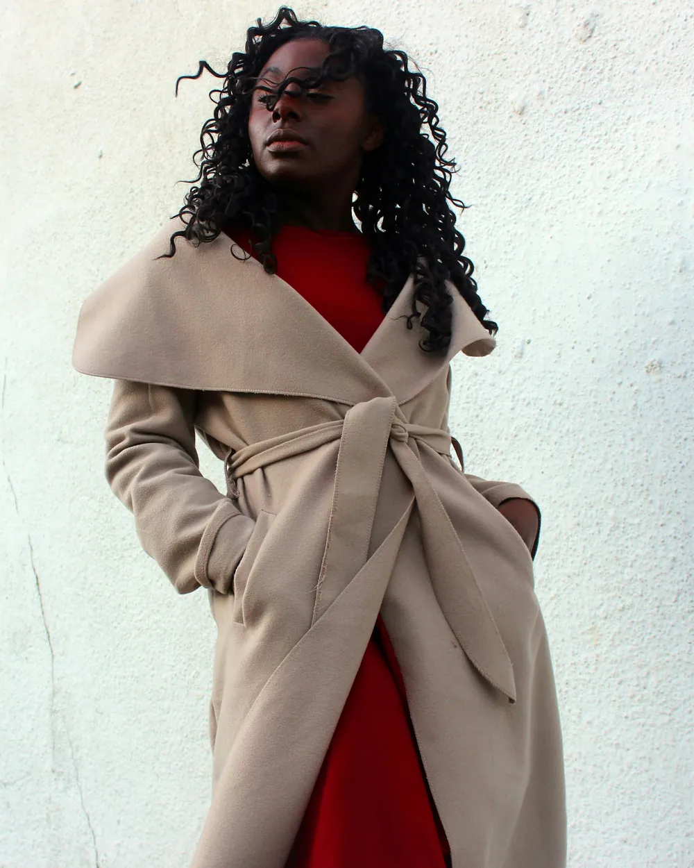 Woman in a camel wrap coat over a red dress against a white textured wall